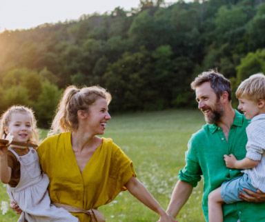 Eine vierköpfige Familie, bestehend aus zwei Erwachsenen und zwei kleinen Kindern, steht lächelnd und Händchen haltend auf einer Wiese mit Bäumen im Hintergrund.