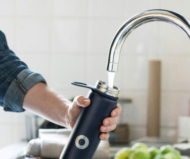 Eine Person füllt eine wiederverwendbare Trinkflasche unter einem Wasserhahn mit Leitungswasser. Im Hintergrund ist eine Küche mit unscharfen Gegenständen zu sehen.