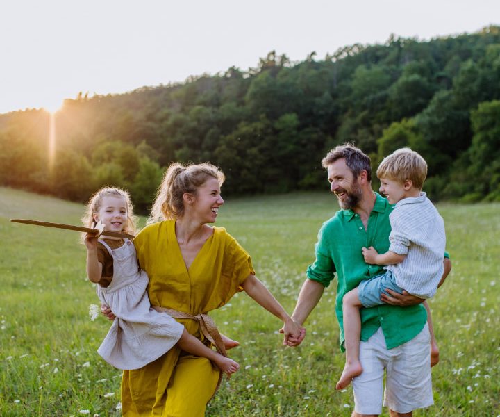 Eine vierköpfige Familie, bestehend aus zwei Erwachsenen und zwei kleinen Kindern, steht lächelnd und Händchen haltend auf einer Wiese mit Bäumen im Hintergrund.