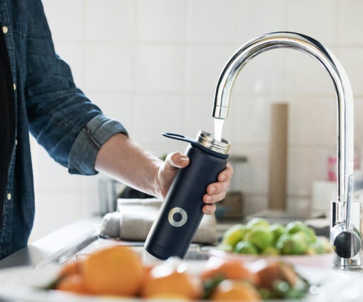 Eine Person füllt eine wiederverwendbare Trinkflasche unter einem Wasserhahn mit Leitungswasser. Im Hintergrund ist eine Küche mit unscharfen Gegenständen zu sehen.