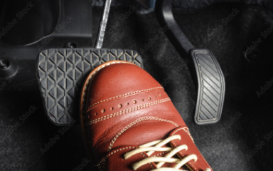 Ein Fuß in braunem Lederschuh tritt auf das Bremspedal eines Autos. Daneben sind Gas- und Kupplungspedal zu sehen.