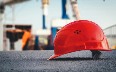 Ein roter Schutzhelm liegt auf dem Boden einer Baustelle, im Hintergrund sind unscharf Baukräne und Gerüste zu sehen.
