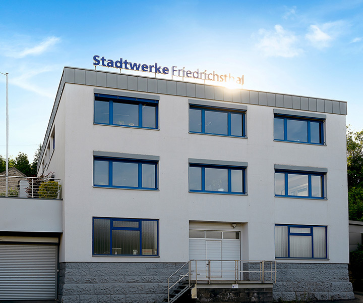 Ein dreistöckiges weißes Gebäude mit blau umrandeten Fenstern und einem Schild auf dem Dach mit der Aufschrift „Stadtwerke Friedrichsthal” vor blauem Himmel.